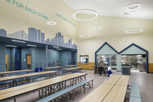 Rows of cafeteria tables fill a large room painted in pale yellow and teal.