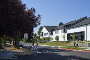 A white school building is lit by sunlight. Across the street, children enter a crosswalk toward the school from the surrounding neighborhood.