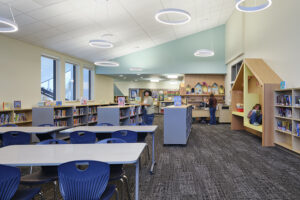 A small school library room is lined with bookshelves around the perimeter with small tables in the foreground.