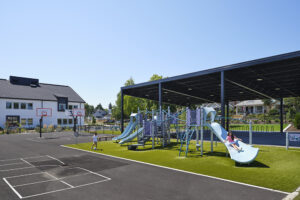 Children play on a bright blue play structure. A large open-sided play shed is visible behind them, and a white school is partially visible on the left side of the image.