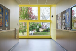 At the end of a hallway, a large yellow-framed window gives a view to trees and houses outdoors.