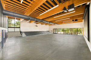 The inside of a large commons space. Windows are on the far wall. The floor is concrete and there are large wooden beams crossing the ceiling. Bleacher seating is set at the opposite wall.