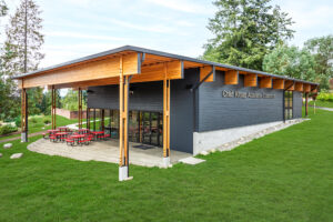 A single-story rectangular building with dark walls and large wooden overhangs. Red patio tables are arranged underneath the overhang.