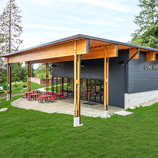 A single-story rectangular building with dark walls and large wooden overhangs. Red patio tables are arranged underneath the overhang.