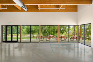 The inside of a large commons space with a concrete floor, wraparound windows, white walls above, and wood beams visible at the ceiling.