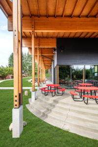 A dark-walled rectangular building seen from just under the edge of its large wooden overhanging roof. Wooden beams and columns are predominant over the covered patio, which has several red patio tables on it.