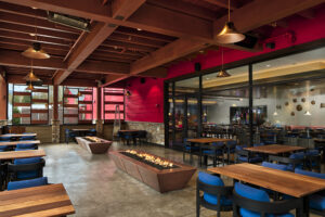 An outdoor patio dining area with low stone fire pits, a wood-beam ceiling and a wall with red siding and large windows viewing the interior seating area.
