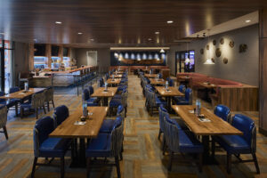 A warmly lit dining room with rows of wood tables surrounded by blue dining chairs.