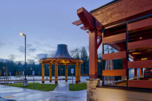 The wooden corner of the casino's outdoor patio is visible with protruding wood beams. In the background, a round salmon bake structure has six wood columns supporting a conical cedar roof.