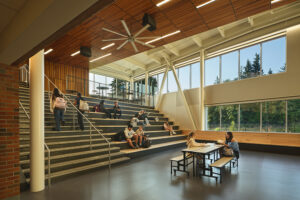A large room with windows on the back wall has a wide staircase on the left side. Students are seated across different parts of the staircase, and two more students sit at a table near the base of the stairs.