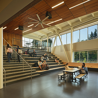 A large room with windows on the back wall has a wide staircase on the left side. Students are seated across different parts of the staircase, and two more students sit at a table near the base of the stairs.