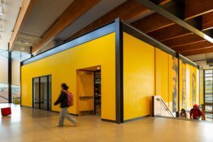 A high school student walks in front of a yellow classroom within the larger space.