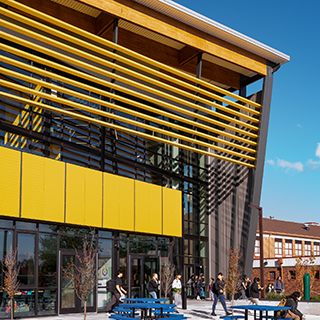 A dramatic black and yellow building juts upward at a slight angle. Students walk in and out from the entry at the bottom center of the building.