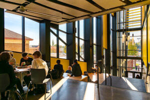 Several high school students gather at the top of a staircase. Windows make up the left side wall separated by steel columns and yellow areas of wall.