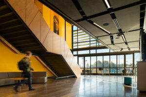 A staircase with a geometric wood pattern along the outside railing descends in front of a tall yellow wall. A large windowed wall lights the background.