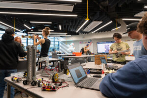 High school students work together in a large, windowed classroom. Laptops and robot parts fill the table space in front of them.