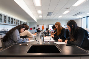 Several high school girls sit around a lab table working on schoolwork. A lab sink is in the foreground.