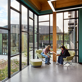 Two high school students sit and talk in front of large, floor-to-ceiling windows. A wood column is in the center of the image just behind them, with more wood beams visible above.
