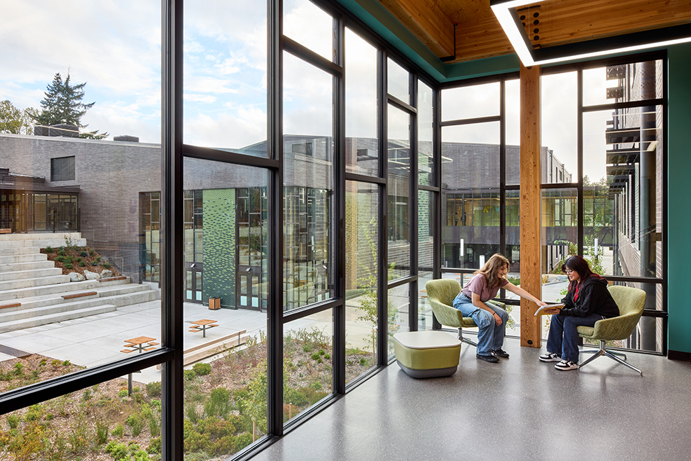 Two high school students sit and talk in front of large, floor-to-ceiling windows. A wood column is in the center of the image just behind them, with more wood beams visible above.