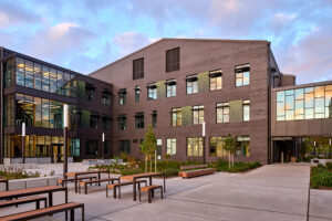 A courtyard with wooden benches is framed by a large brick building with three stories of windows. At the right side, a pedestrian sky bridge is visible.