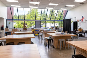 A classroom with wooden tables and stools. The far wall is a tall, angled window.