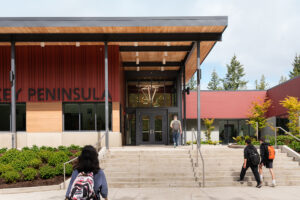 A middle school student walks across a crosswalk toward a school building which is red with wood accents and black steel columns.