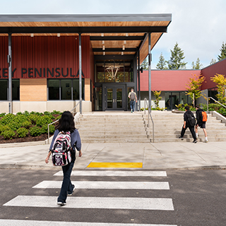 A middle school student walks across a crosswalk toward a school building which is red with wood accents and black steel columns.