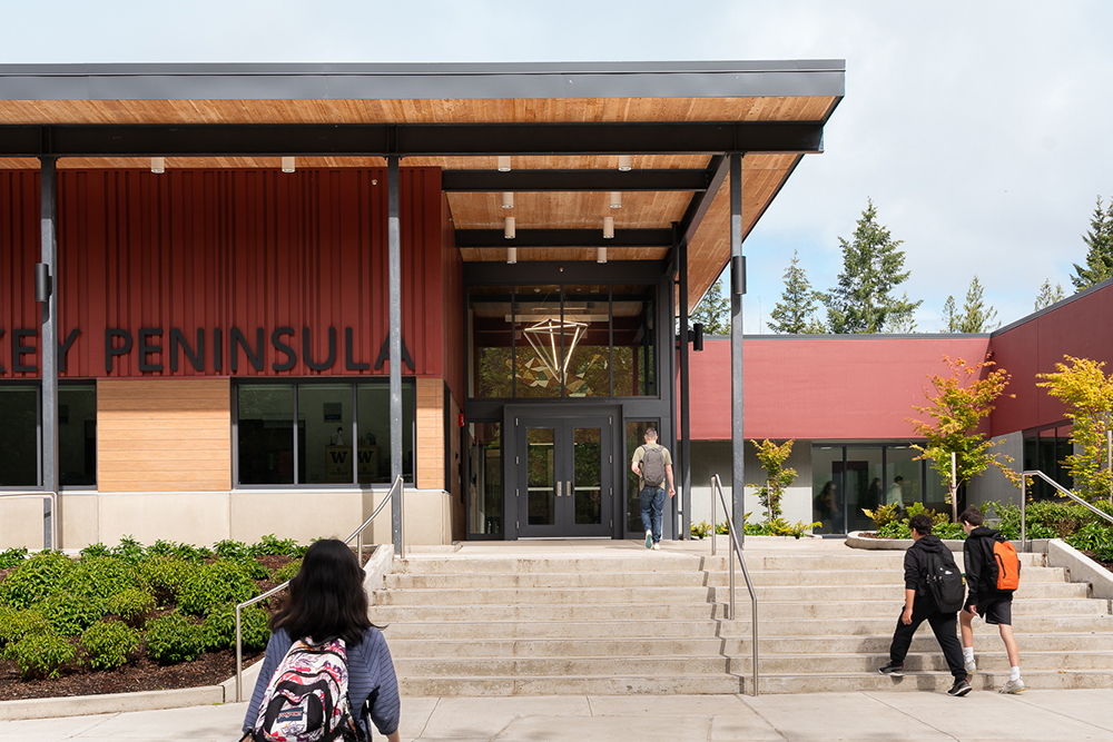 A middle school student walks across a crosswalk toward a school building which is red with wood accents and black steel columns.