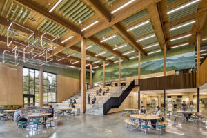 High school students work at round tables arranged in a large two-story commons space. Wood ceilings, beams, and columns create a warm and inviting space.