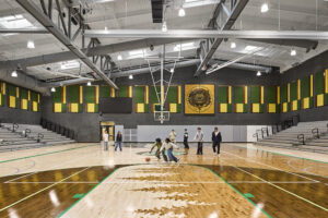 High school students play basketball inside a large gymnasium with a wooden floor, dark walls with green and gold banners, and a white metal ceiling.