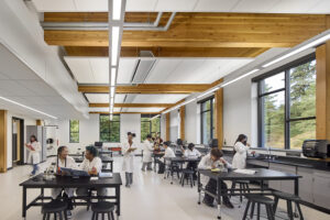 High school students dressed in white lab coats work on projects at rows of black tables. They are in a mostly white room with large wood beams at the ceiling and tall windows along the right side wall.