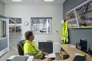 A man wearing a bright yellow high-vis shirt sits at a desk and looks at a monitor on the wall with images of various locations on it.