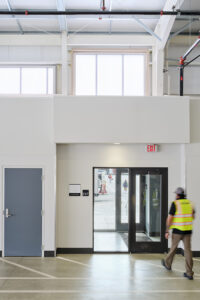 A man wearing a high-vis yellow vest walks toward a door inside a bus garage.