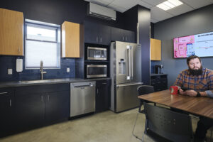 A blue-painted staff kitchen with yellow accents. A staff member in a plaid shirt sits at the table on the right side.
