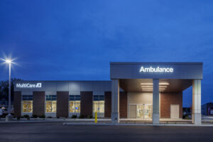 The side of an emergency department building with brick walls and white accents. A large canopy labeled "Ambulance" juts out from the building on the right side of the photo.