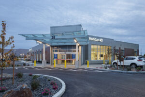 A single-story emergency department building with brick walls and white-painted accents. A broad glass canopy is cantilevered over the entrance. The sign reads "MultiCare."