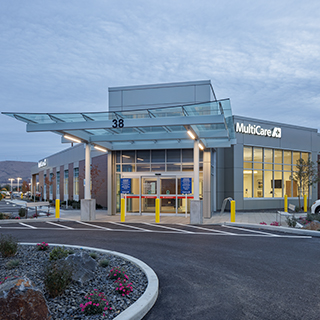 A single-story emergency department building with brick walls and white-painted accents. A broad glass canopy is cantilevered over the entrance. The sign reads "MultiCare."