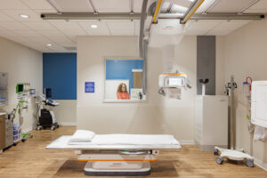 A hospital room with a small flat bed in the center and imaging equipment hung from the ceiling. A person dressed in orange is visible through a window in the back wall.