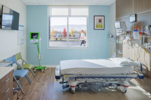 A small hospital patient room with a bed and various medical equipment. A window on the far wall frames distant autumn trees and hills.