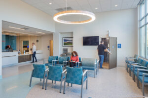 A small waiting room in an emergency department. Six blue chairs are lined up in the foreground, one of them occupied by a person. In the background, there is a man waiting to talk to someone behind a desk. Another man walks through a door into a restroom at the far right.