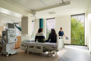 Two people sit on a hospital bed with their backs facing the camera. A nurse passes between them and the wall which has two tall windows.