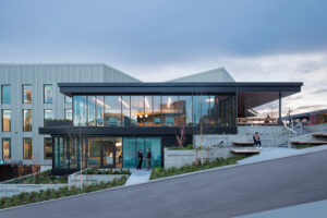 A two-story clinic building with full-height glass walls sits on a steel hill. Several people are sitting on concrete terraces in front of the building.