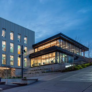 A two-story clinic building rests on a hillside beside an adjacent four-story rectangular building. The windows glow warmly in the twilight.