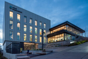 A two-story clinic building rests on a hillside beside an adjacent four-story rectangular building. The windows glow warmly in the twilight.