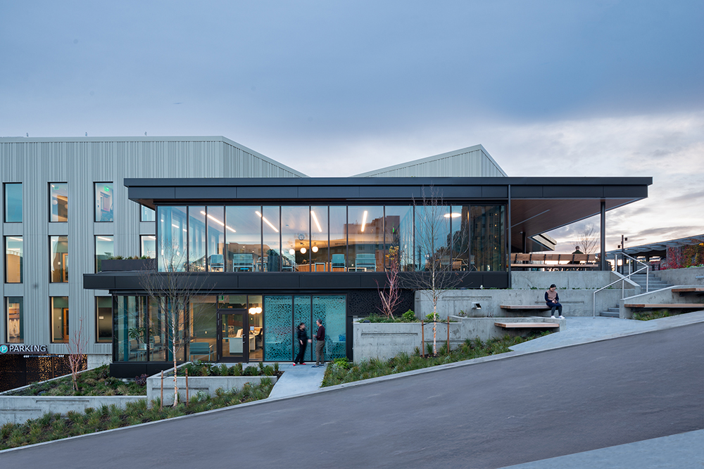 A two-story clinic building with full-height glass walls sits on a steel hill. Several people are sitting on concrete terraces in front of the building.