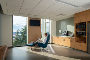 A physician in a white coat rests in a blue chair, illuminated by afternoon light from full-height windows. A staff kitchen counter is behind him.
