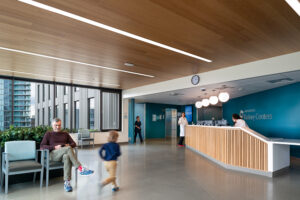 A man sits in a chair in a large waiting room. Large windows on the far wall show a city skyline outside. Several medical workers are near the reception counter to the right. The ceiling is smooth and glossy wood.