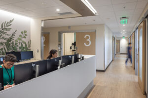 Two nurses work at a row of computers at a nurses's station. In the background is a row of patient rooms.