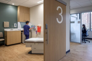 A medical worker in blue scrubs works inside a patient exam room.