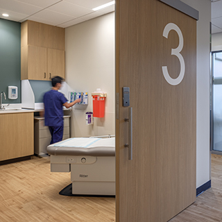 A medical worker in blue scrubs works inside a patient exam room.